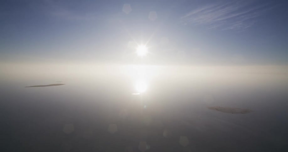 A surreal and symmetrical aerial view captures the bright sun and its reflection over a perfectly calm sea, with two islands visible through the haze