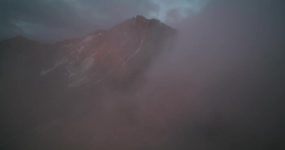 A slow and moody aerial shot pushes through a dense, pink-tinged cloud to reveal a rugged, snow-dusted mountain peak at sunrise