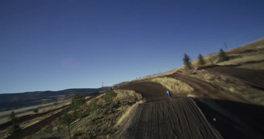 Dynamic FPV drone footage follows a motocross rider performing a big jump in slow motion on a scenic hillside course