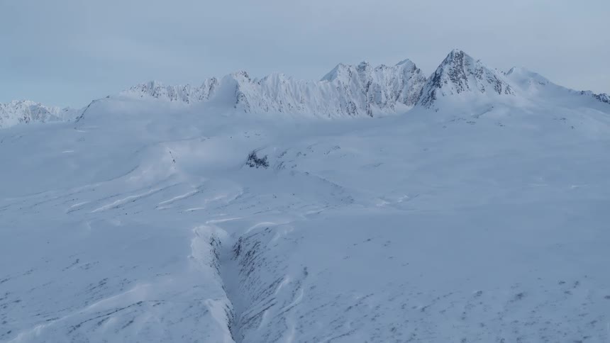 A vast, frozen tundra stretches towards a majestic, snow-covered mountain range under an overcast sky