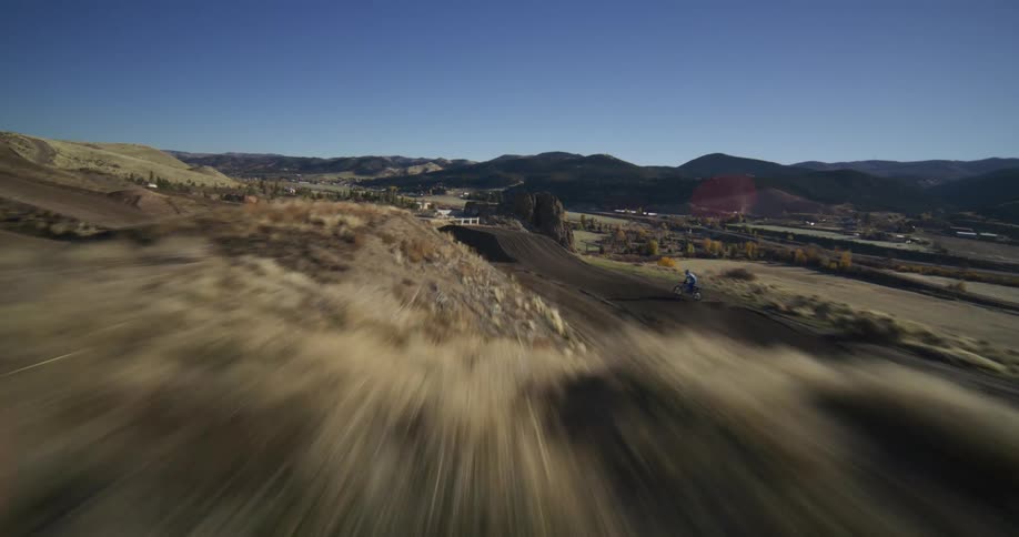 An FPV drone captures a motocross rider in slow motion as they speed along a dirt track and launch off a large jump into the air