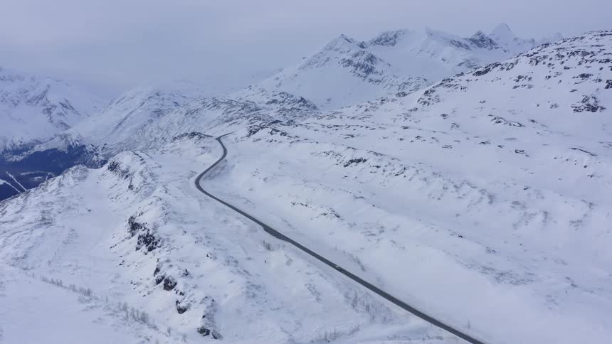 An aerial view shows a lone car driving down a winding road through a majestic, snow-covered mountain range in winter
