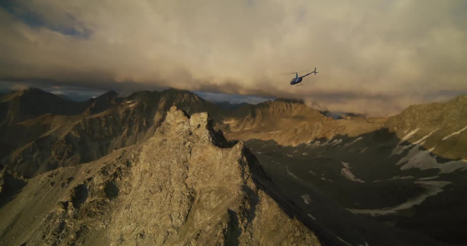 A dramatic aerial shot follows a helicopter as it flies over towering, rugged mountain peaks during a beautiful sunset