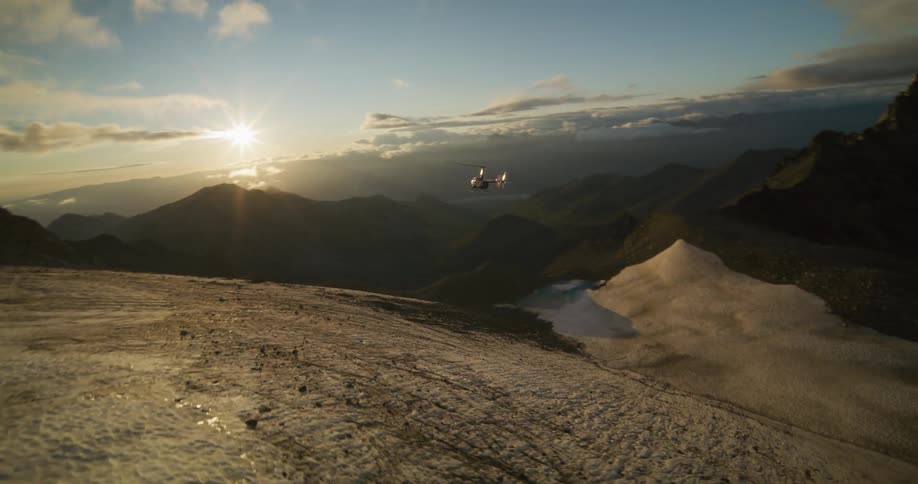 A helicopter flies away from a snowy glacier over a vast mountain range as the sun rises, creating a dramatic silhouette