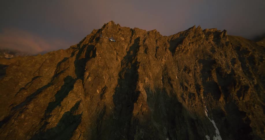 An aerial view shows a helicopter flying towards a rugged, sun-drenched mountain peak at sunset