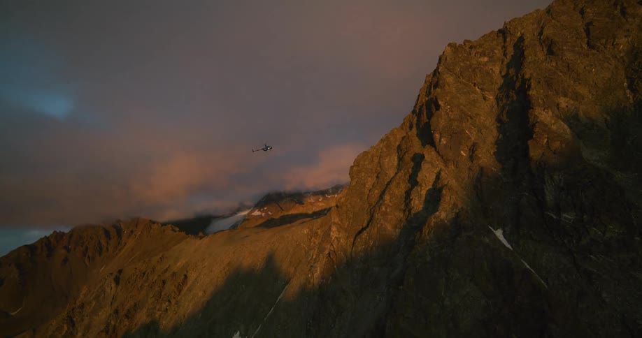 A lone helicopter soars past a magnificent, rugged mountain peak bathed in the golden light of sunset