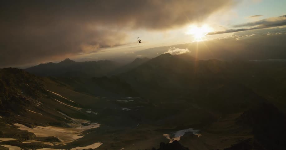 A stunning aerial view shows a helicopter soaring over a majestic, shadowed mountain range during a golden sunset
