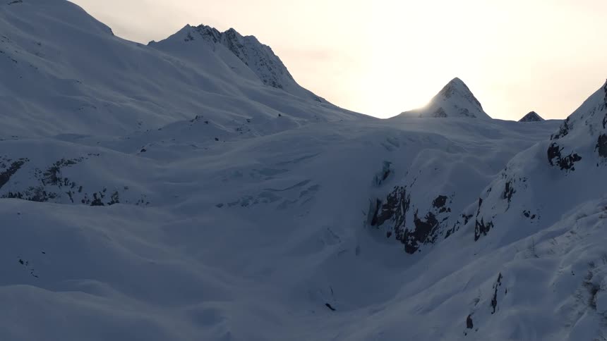 A slow, majestic drone shot pushes through a vast, snow-covered mountain valley as the sun rises over a distant peak
