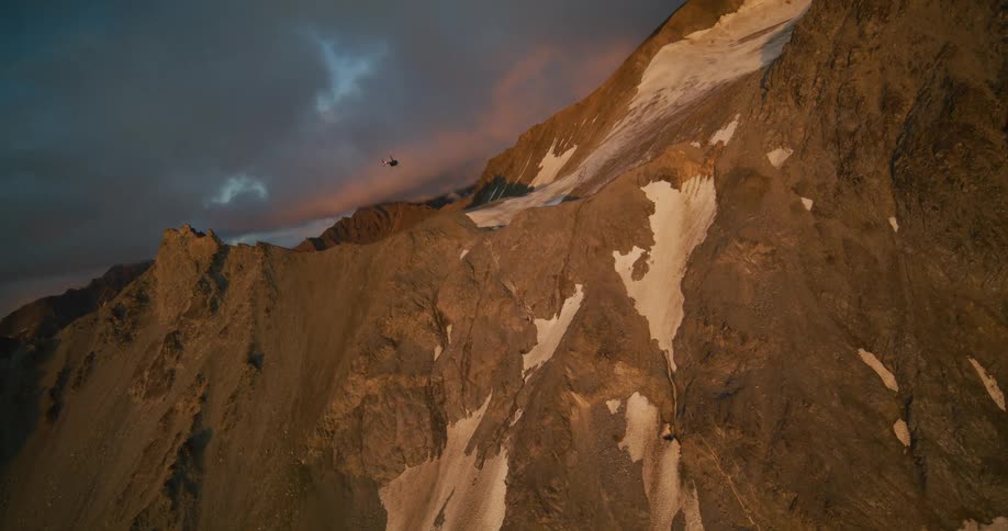A helicopter flies away from a rugged, snow-dusted mountain range under a dramatic sunset sky