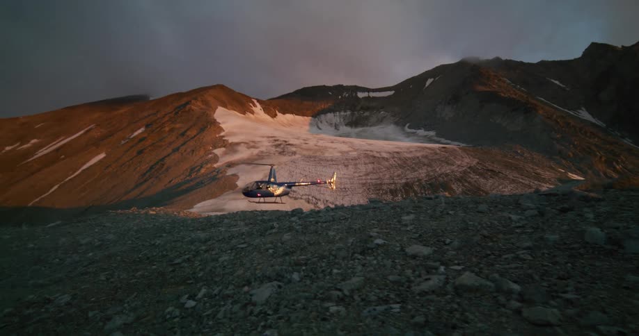 A helicopter hovers above a rocky mountain pass next to a glacier during a dramatic and beautiful sunset