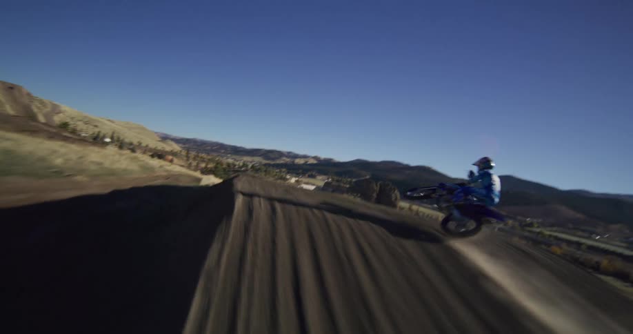 A dynamic FPV drone chases a motocross rider in slow motion as they launch off a huge dirt ramp on a sunny day