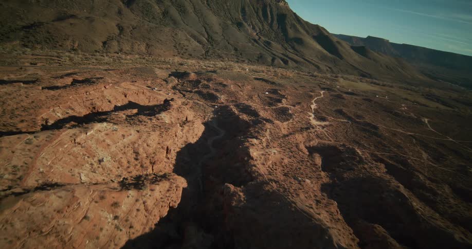 An epic aerial reveal shot flies backwards over rugged red rock formations to show a vast desert valley at the base of a large mesa
