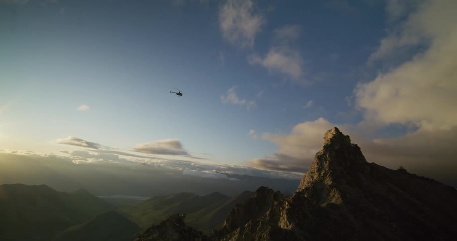 A silhouetted helicopter flies across a stunning mountain range, passing rugged peaks during a beautiful sunrise