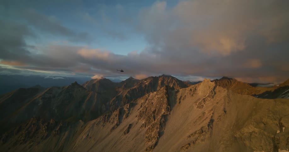 A helicopter soars over a rugged mountain range, its peaks illuminated by the warm light of sunrise under a dramatic, cloudy sky
