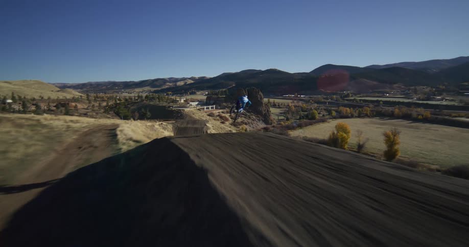 A high-speed FPV drone chases a motocross rider as they launch off a massive jump on a dirt track in a beautiful mountain landscape
