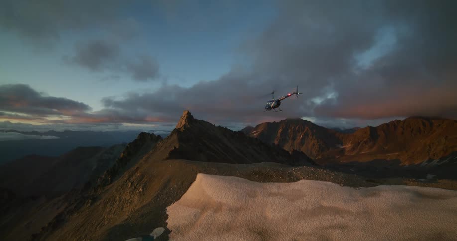 A slow-motion wide shot of a helicopter flying over a rugged mountain range and landing on a peak at sunset
