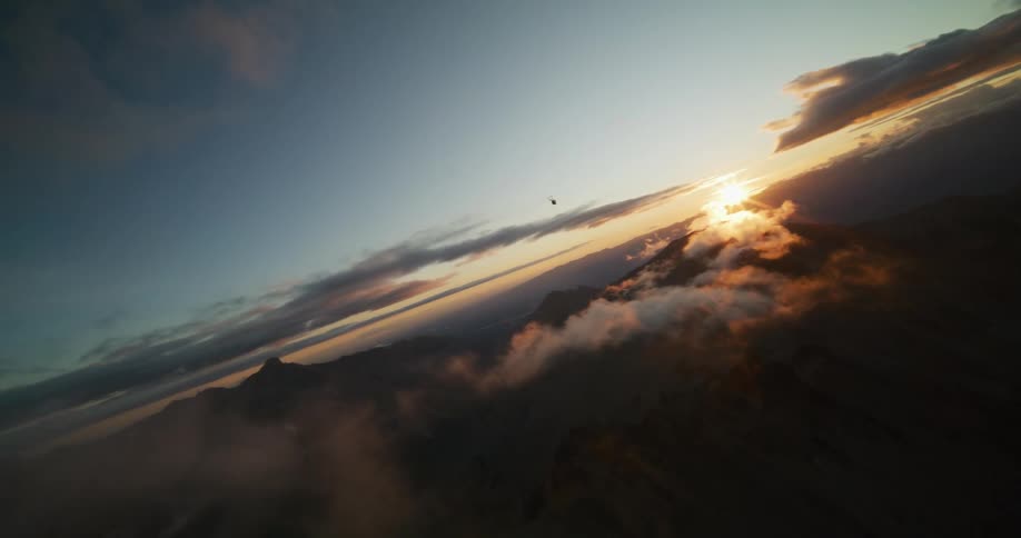 A stunning canted-angle aerial shot captures a helicopter flying over a vast mountain range during a breathtaking sunrise