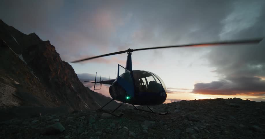 A helicopter with its rotor blades spinning sits on a rocky mountain ridge as the sun rises over the horizon