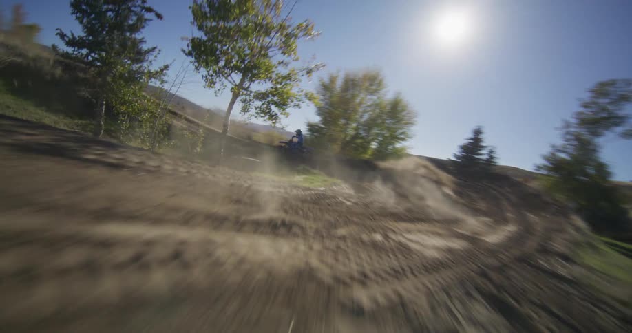 An FPV drone follows a motocross rider as they speed around a dusty dirt track and launch off a jump on a sunny day
