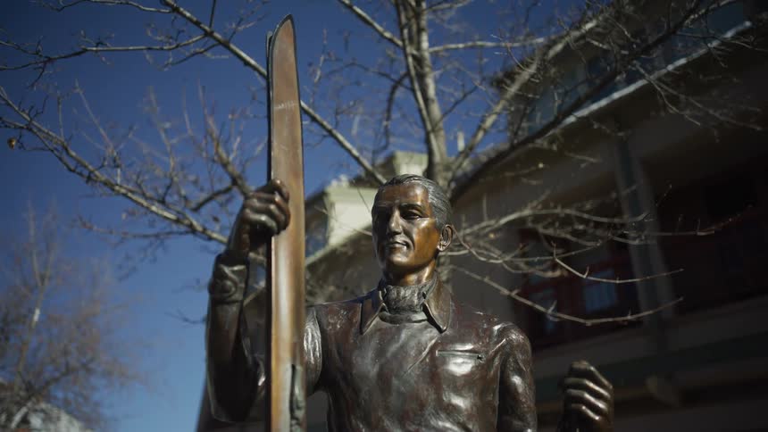 A low-angle shot captures a bronze statue of a man holding a ski against a clear blue sky