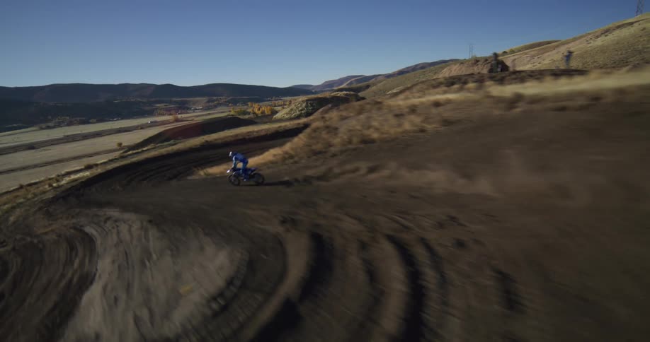 Exhilarating FPV drone footage follows a motocross rider speeding around a dusty dirt track on a sunny hillside