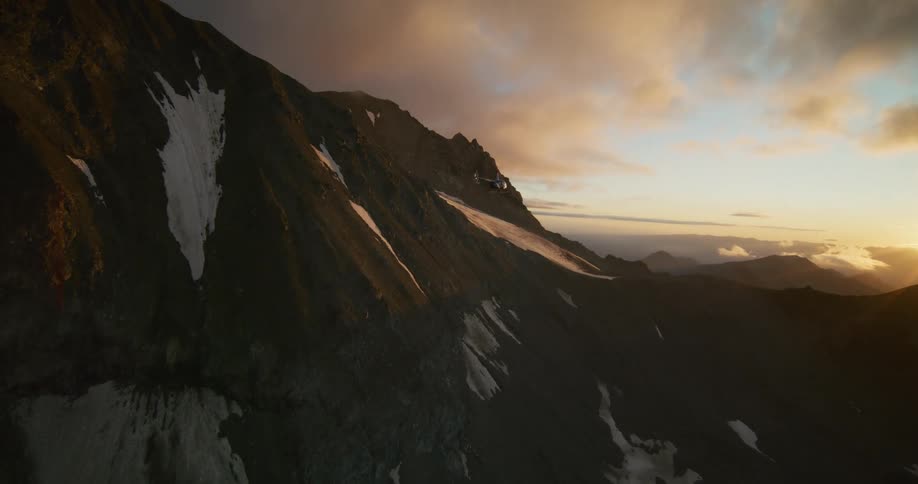 A helicopter soars across a rugged, snow-dusted mountain landscape as the sun sets in a dramatic, cloudy sky