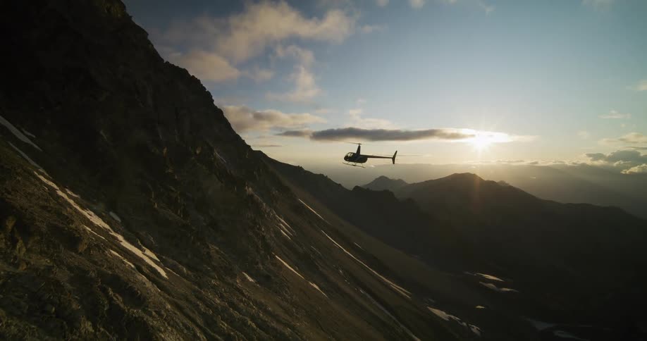 A silhouetted helicopter flies slowly over a stunning mountain valley during a beautiful sunset