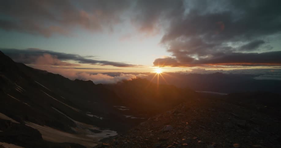 A golden sun rises over a vast and rocky mountain range under a dramatic, cloudy sky