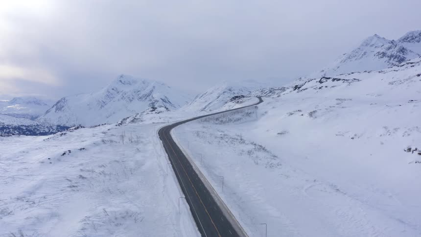 Aerial view of a desolate highway winding through a vast, snow-covered mountain landscape in winter