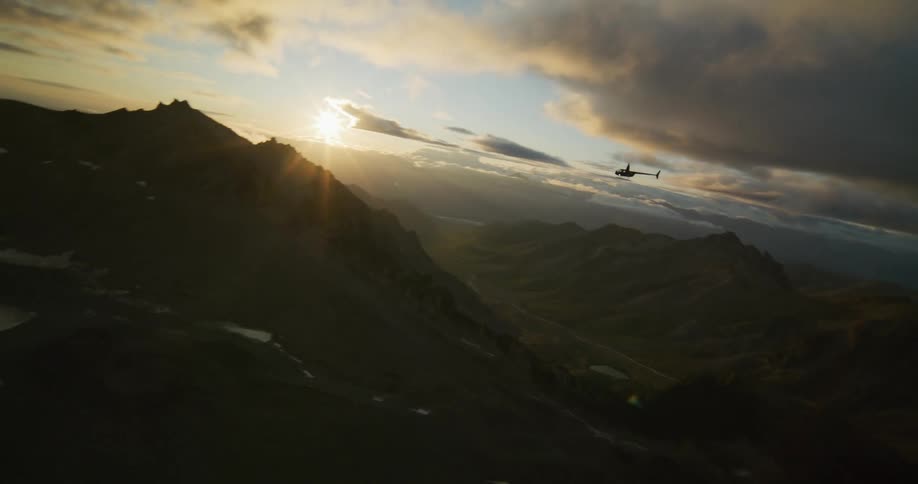 A helicopter flies through a majestic, shadowed mountain range into the beautiful, bright sunset