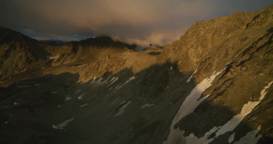 Aerial view of a helicopter flying over a dramatic, golden-lit mountain range before landing on a snowy summit at sunset
