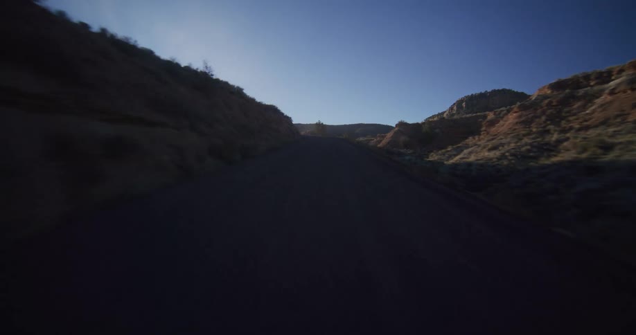 A point-of-view shot driving along a winding desert road through a shadowy canyon toward the bright morning sun