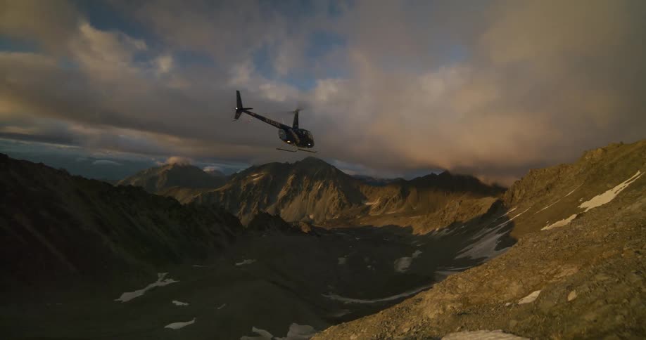 A helicopter soars through a vast, snow-dusted mountain range, landing on a sunlit ridge at golden hour