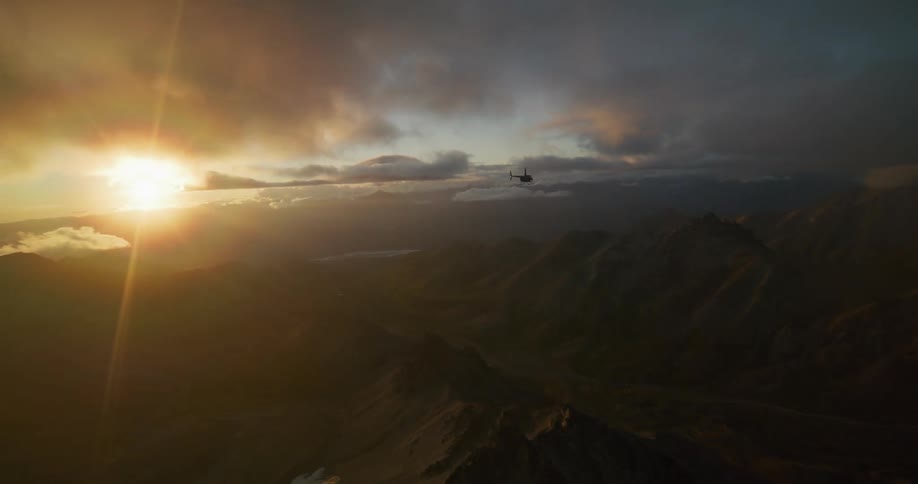 A majestic aerial view shows a helicopter soaring over a rugged mountain range as the golden sun rises through dramatic clouds