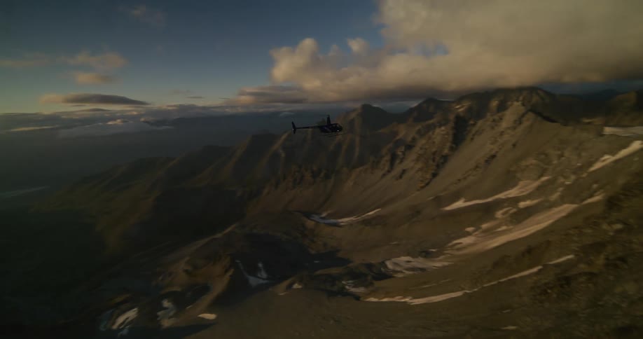 A helicopter flies over a stunningly vast mountain range, silhouetted against the rising sun