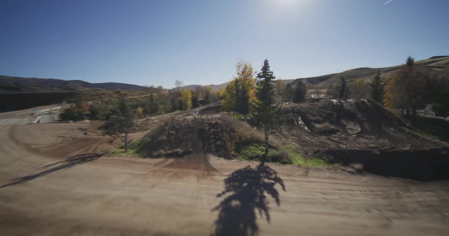 A dynamic FPV drone captures a thrilling slow-motion shot of a motocross rider launching off a large dirt jump on a sunny day