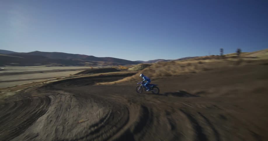 A dynamic tracking shot follows a motocross rider in blue gear as they power through a dusty turn on a winding hillside track on a sunny day