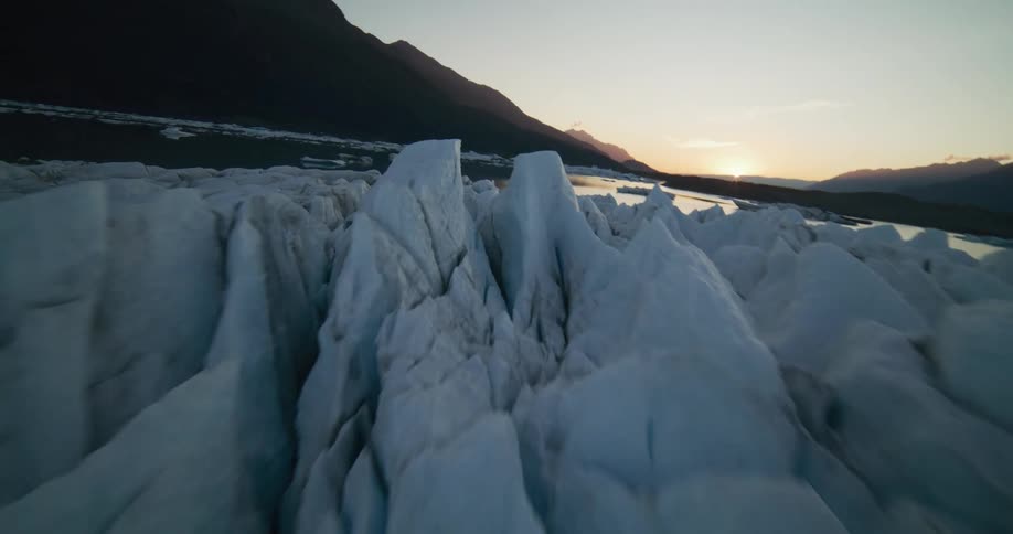 An FPV drone soars closely over the cracked and jagged surface of a massive glacier towards a mountain lake at sunset