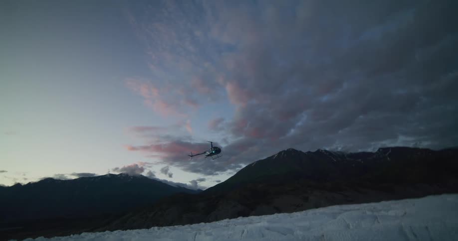 A helicopter flies slowly towards the camera over a vast snowy glacier at twilight against a dramatic cloudy sky