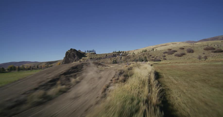 Dynamic slow-motion tracking shot of several motocross riders performing massive jumps on a rural dirt track under a clear blue sky