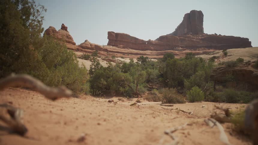 A low-angle shot from a sandy desert wash reveals large red rock formations and buttes under a bright sky