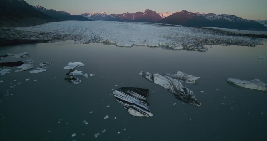 A majestic aerial flight over a serene glacial lake filled with floating icebergs in front of a massive glacier at dawn