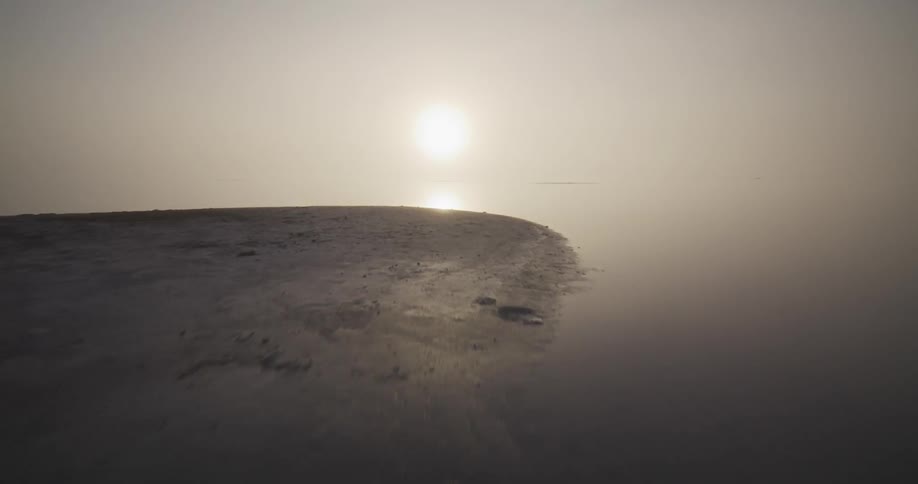 A low aerial shot glides over a misty shoreline and across a calm, reflective lake toward the hazy morning sun