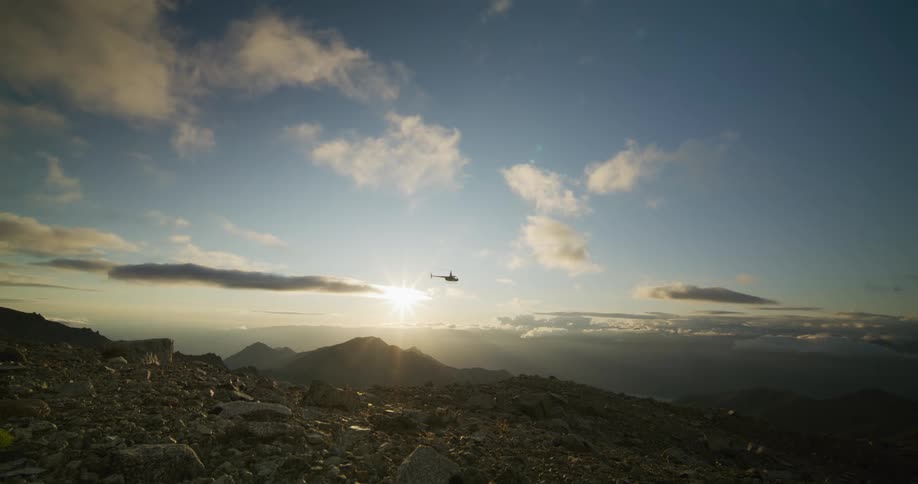 A helicopter flies across a vast, rocky mountain landscape, silhouetted against the beautiful rising sun