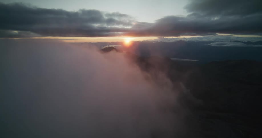 A majestic aerial view emerges from thick clouds, revealing a dramatic mountain range during a beautiful sunset