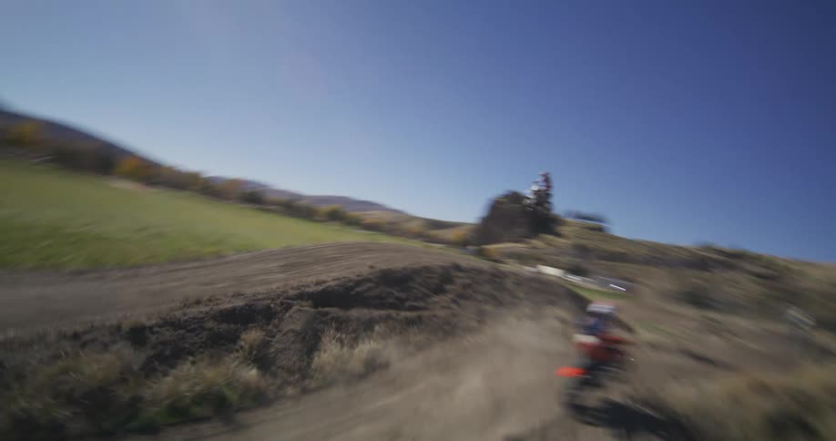 Dynamic aerial footage follows multiple motocross riders as they race and jump over hills on a winding dirt track in the countryside