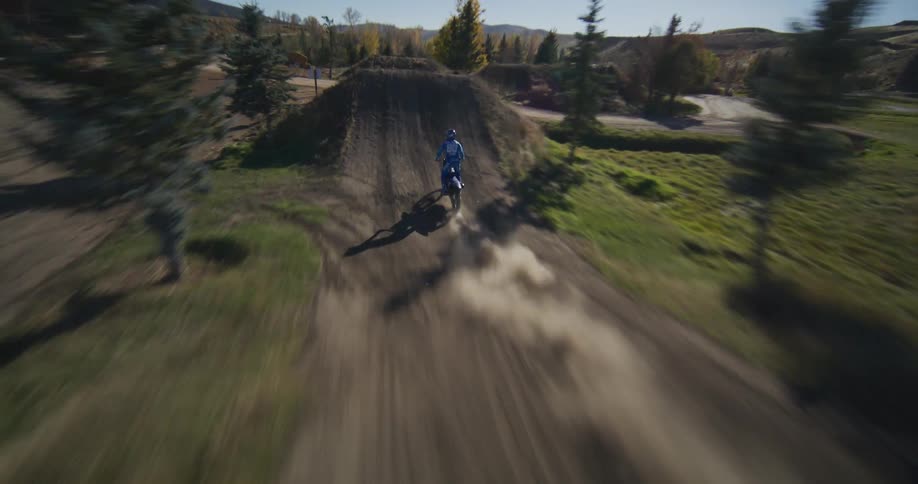 An FPV drone dynamically chases a motocross rider as they speed along a dirt track, launching into the air for spectacular slow-motion jumps and tricks
