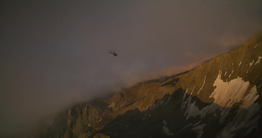 A helicopter flies slowly over a vast, rugged mountain range partially covered in clouds during a dramatic sunset
