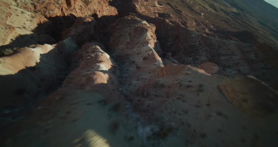 Dynamic FPV drone footage soars over a sunlit desert mountain ridge before diving into a vast, rugged canyon