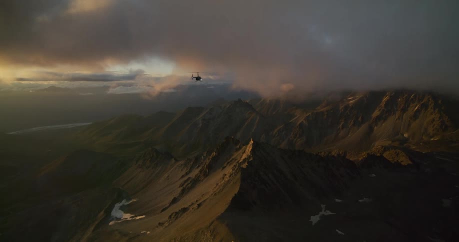 An epic aerial shot follows a helicopter flying over a vast and rugged mountain range shrouded in clouds at golden hour